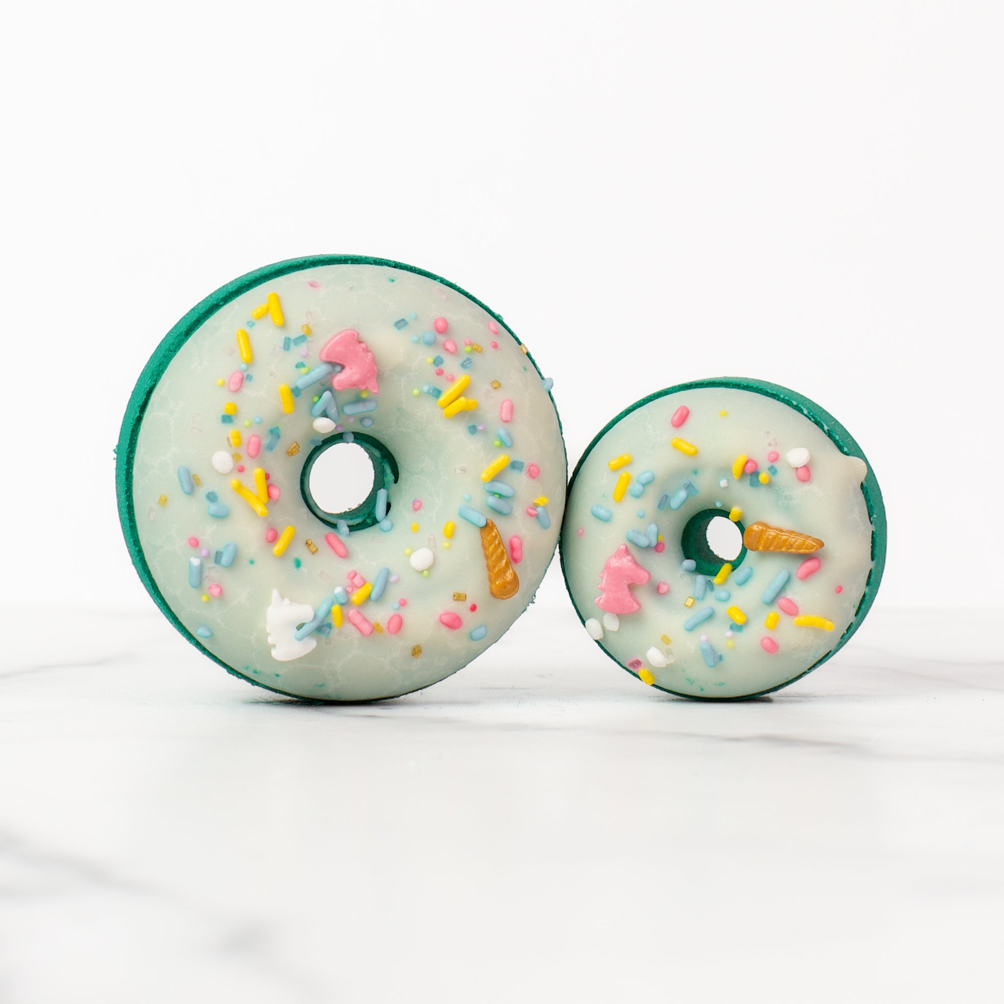 One large and one small donut Volcano bath bombs on a white background. Volcano is a teal bath bomb with a white glaze and pastel unicorn sprinkles.