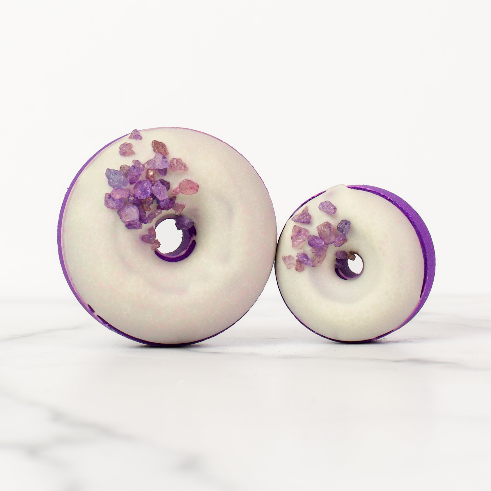 One large and one small donut Moonflower bath bombs on a white background. Moonflower is a dark purple bath bomb with a white glaze and purple gemstone sprinkles.