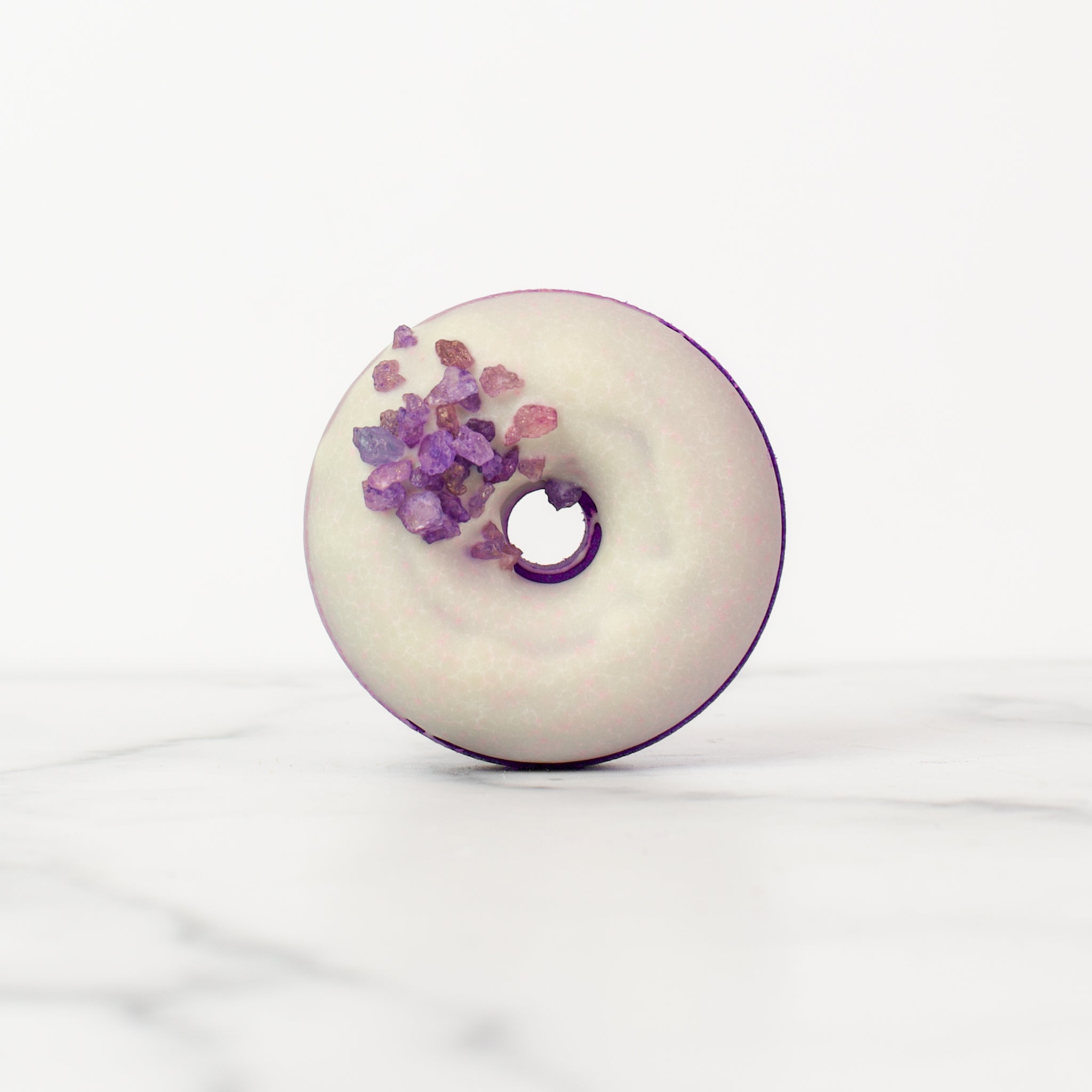 One large donut Moonflower bath bombs on a white background. Moonflower is a dark purple bath bomb with a white glaze and purple gemstone sprinkles.