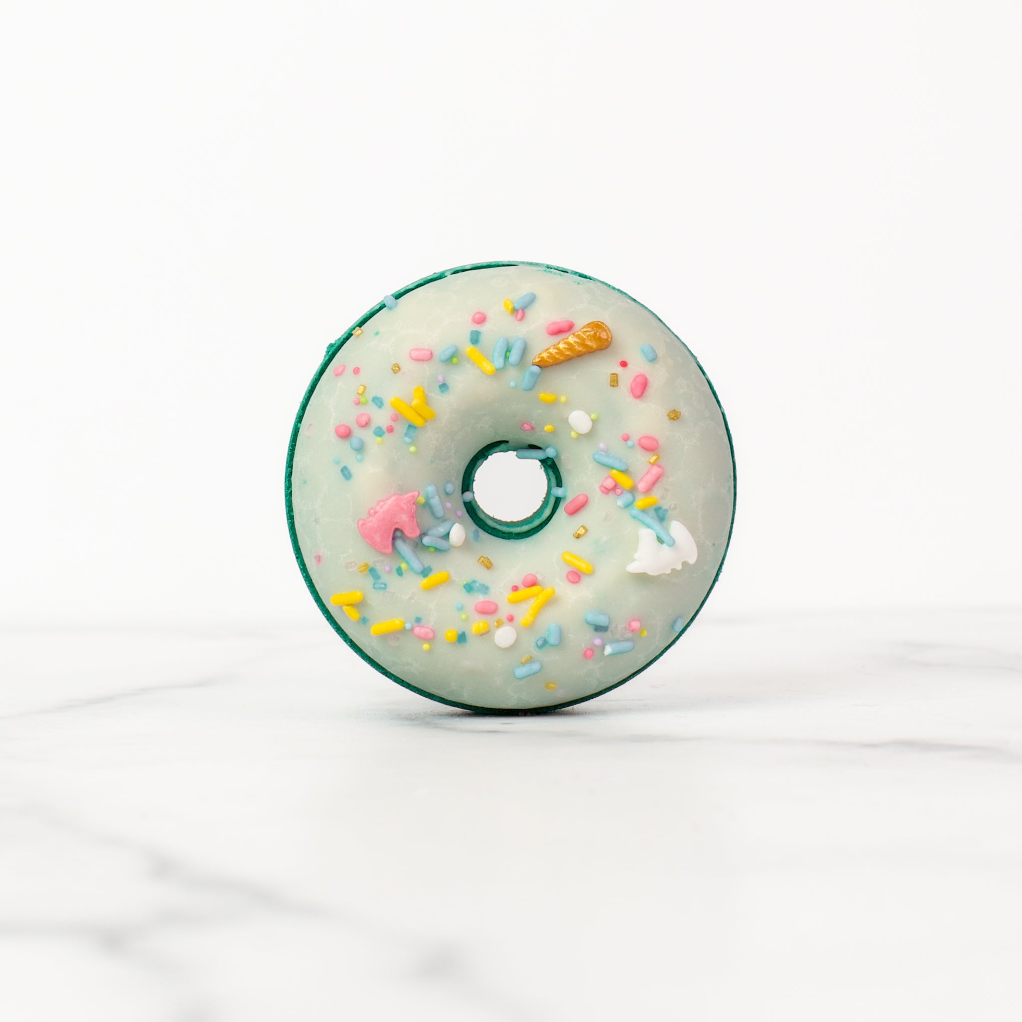 One large donut Volcano bath bombs on a white background. Volcano is a teal bath bomb with a white glaze and pastel unicorn sprinkles.