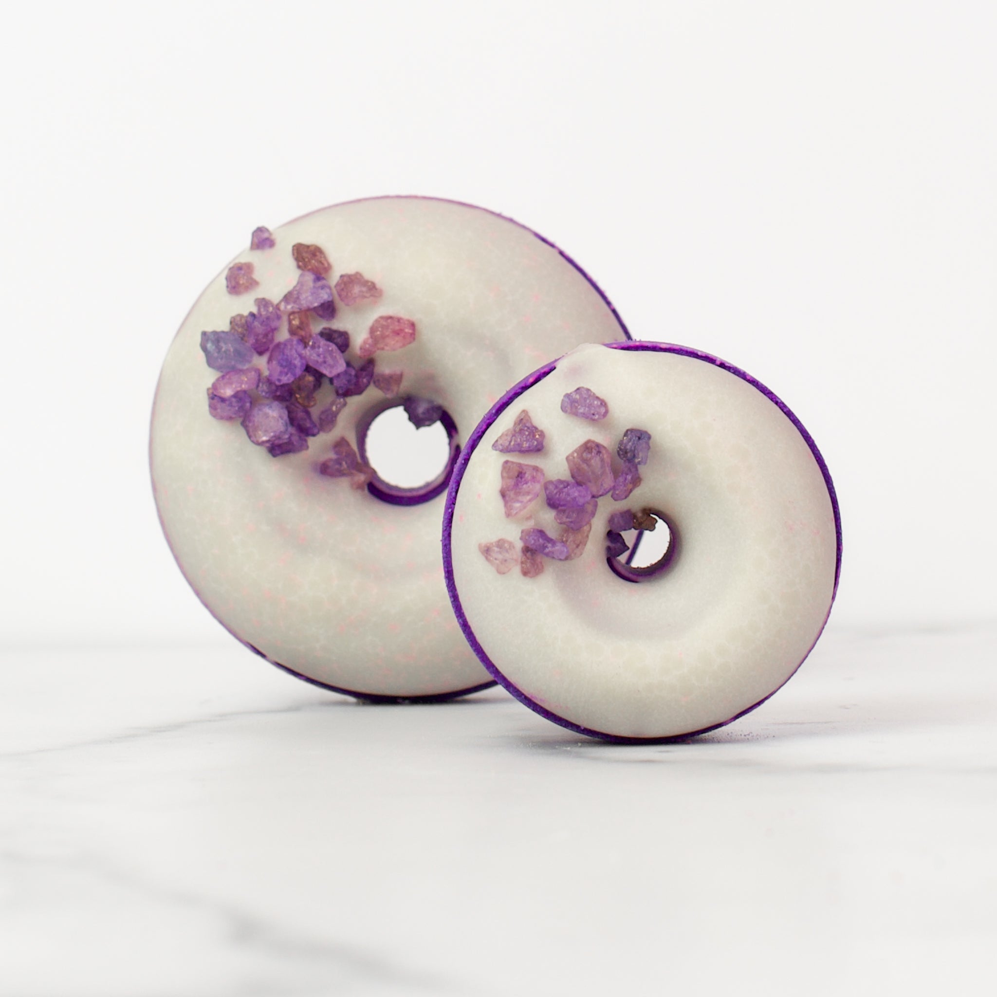 One large and one small donut Moonflower bath bombs on a white background. Moonflower is a dark purple bath bomb with a white glaze and purple gemstone sprinkles.
