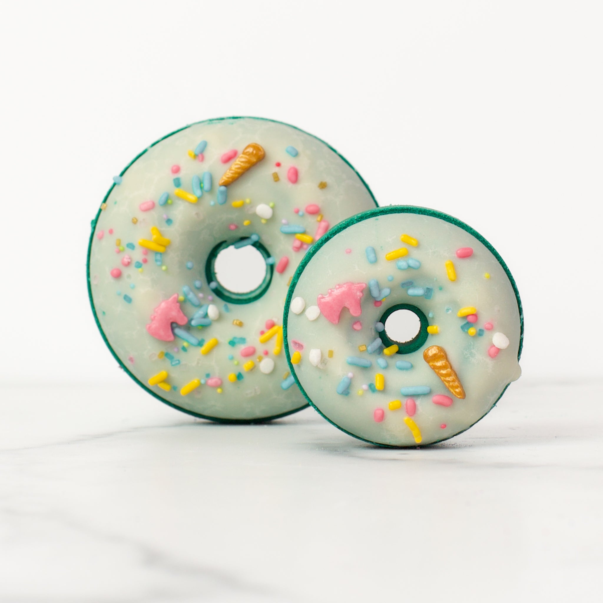 One large and one small donut Volcano bath bombs on a white background. Volcano is a teal bath bomb with a white glaze and pastel unicorn sprinkles.