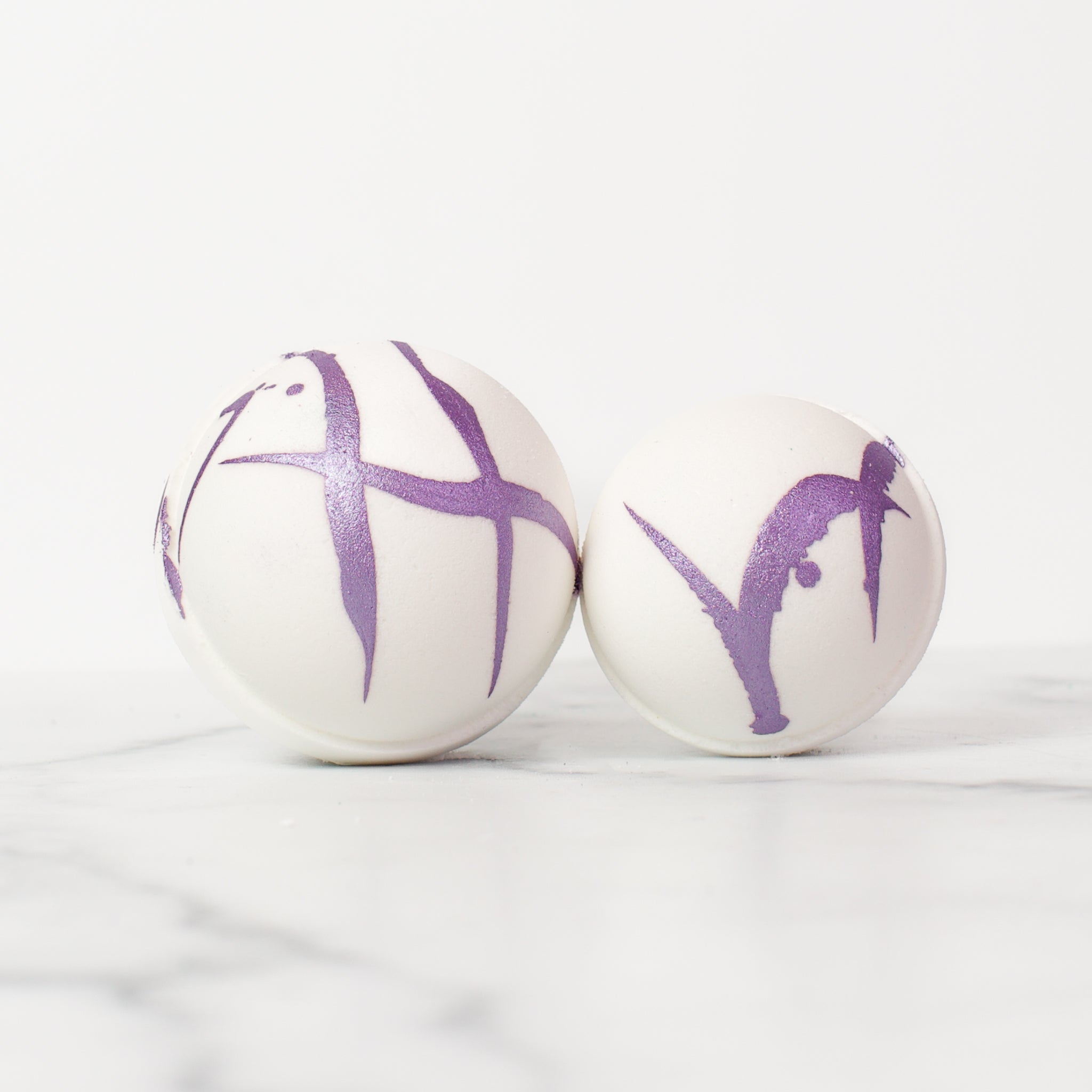 One large and one medium round Lavender bath bombs on a white background. Lavender is a white bath bomb with a light silver-purple splatter design.