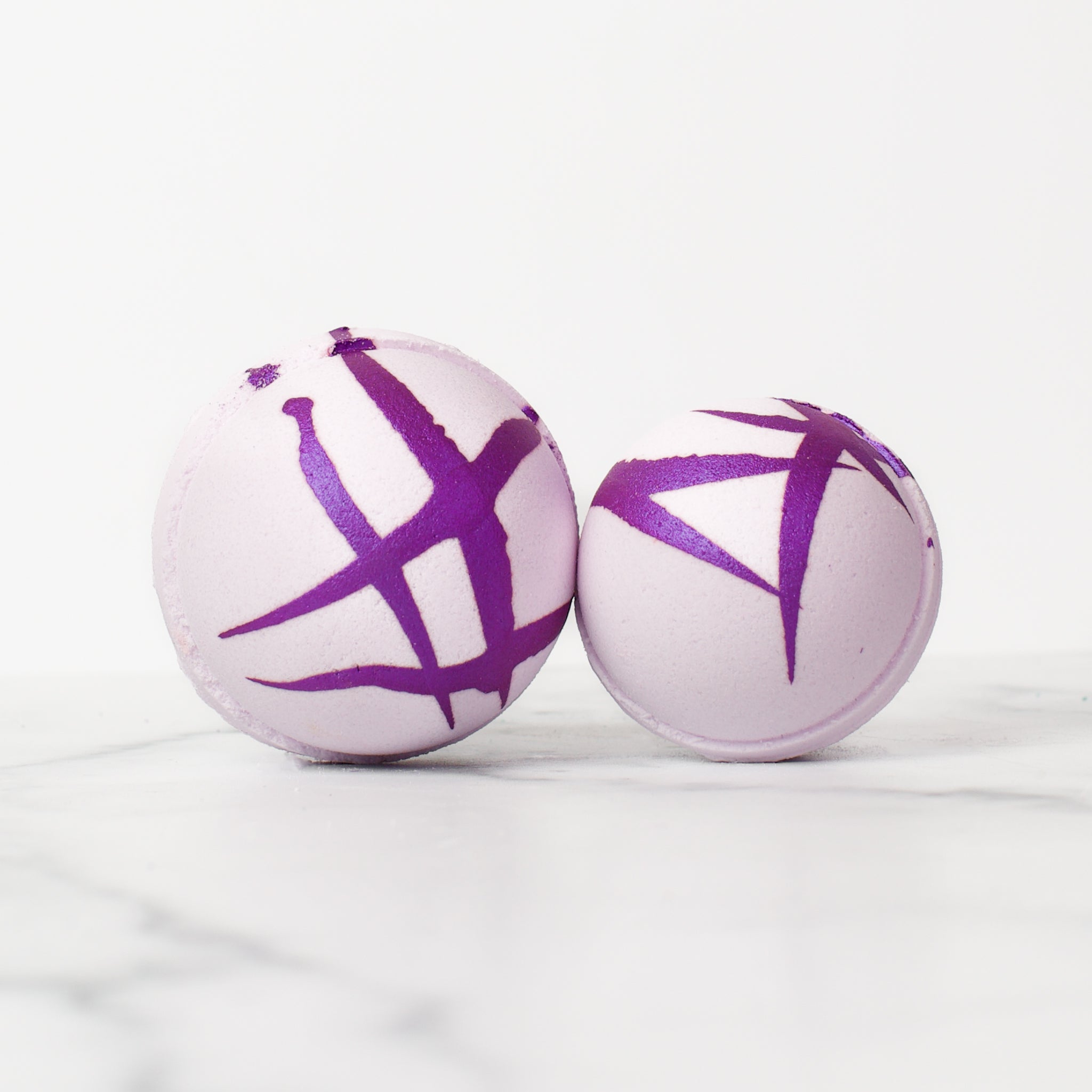 One large and one medium round Lavender & Palmarosa bath bombs on a white background. Lavender & Palmarosa is a light purple bath bomb with a dark purple splatter design.