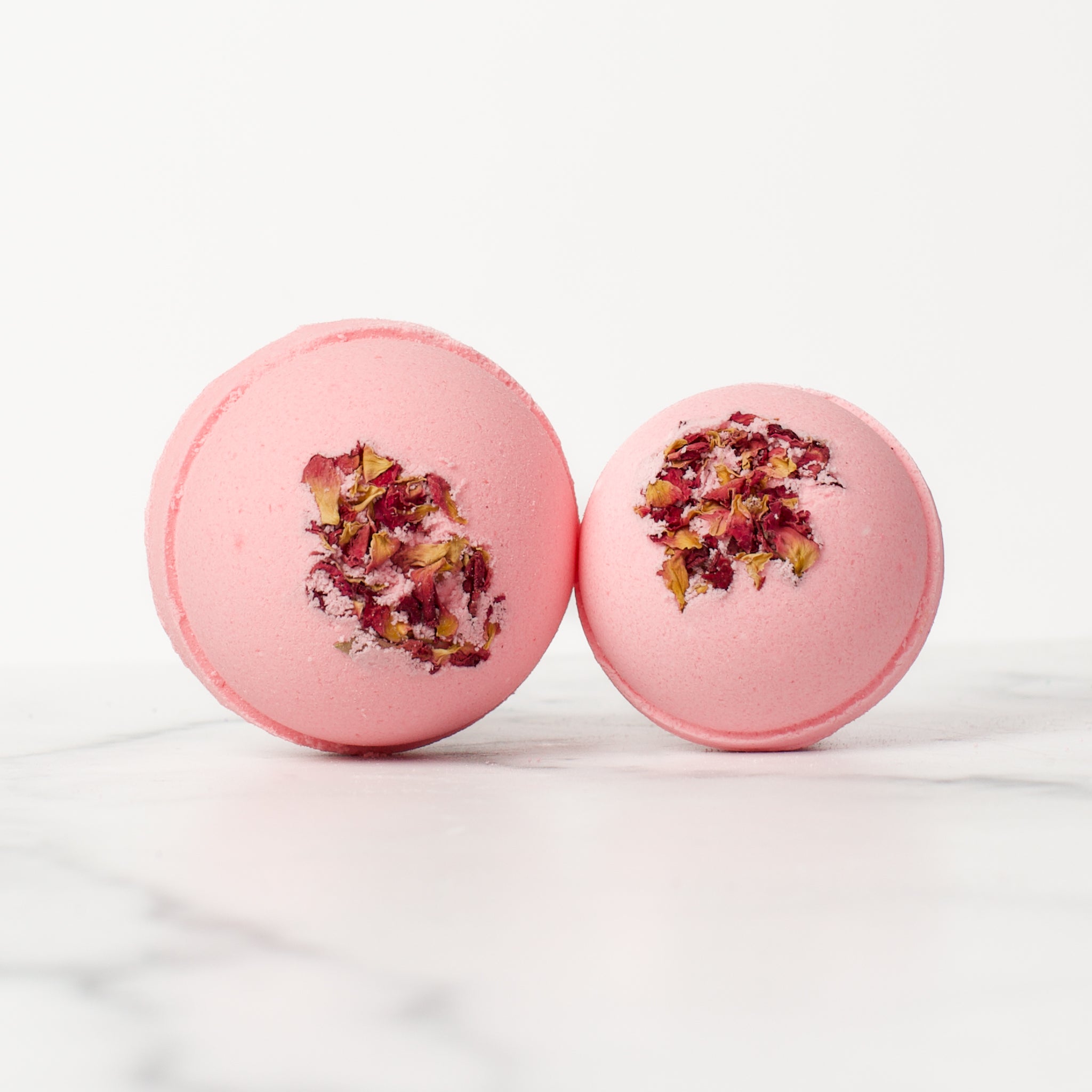 One large and one medium round Lemon Rose bath bombs on a white background. Lemon Rose is a light pink bath bomb with rose petals embedded on the top..