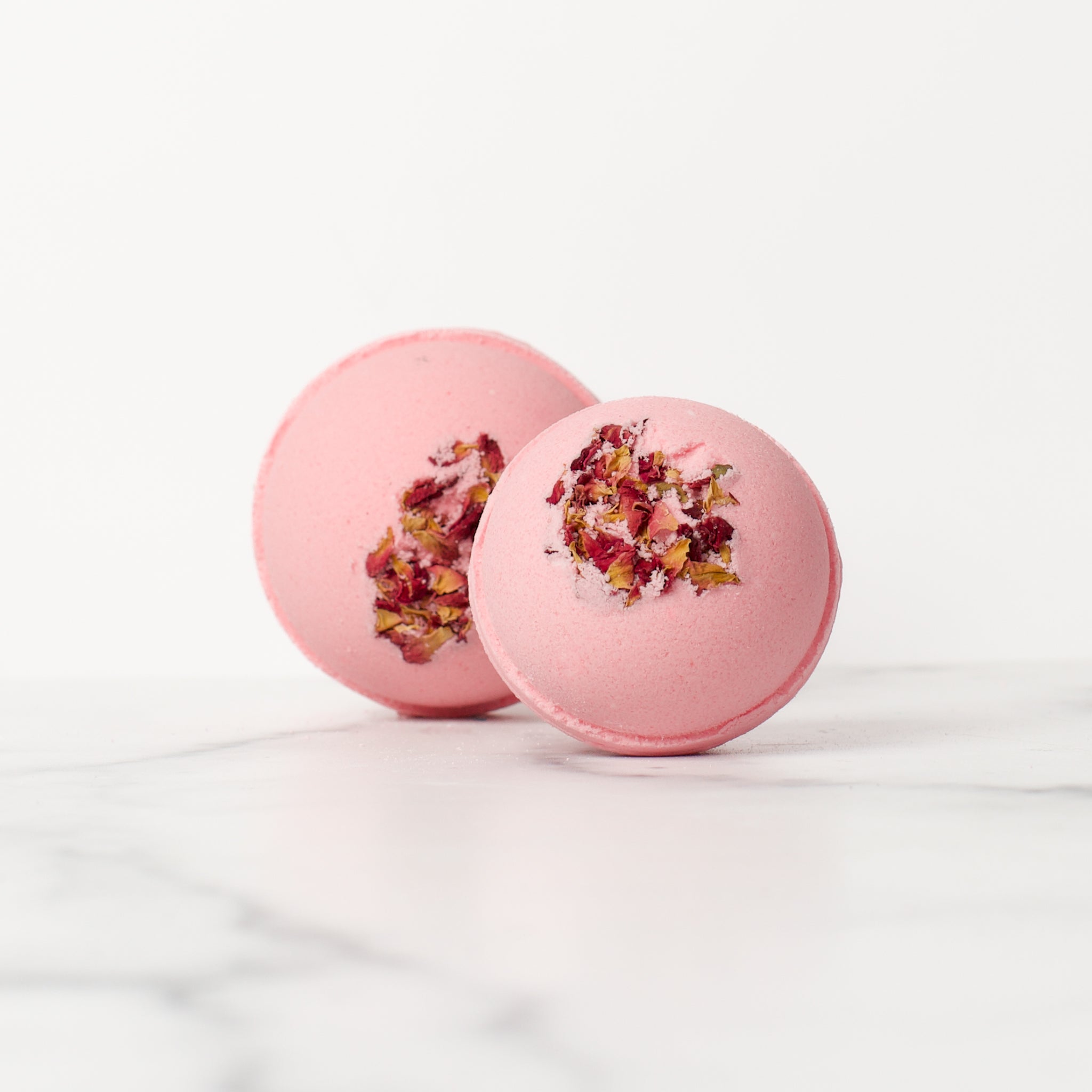 One large and one medium round Lemon Rose bath bombs on a white background. Lemon Rose is a light pink bath bomb with rose petals embedded on the top..