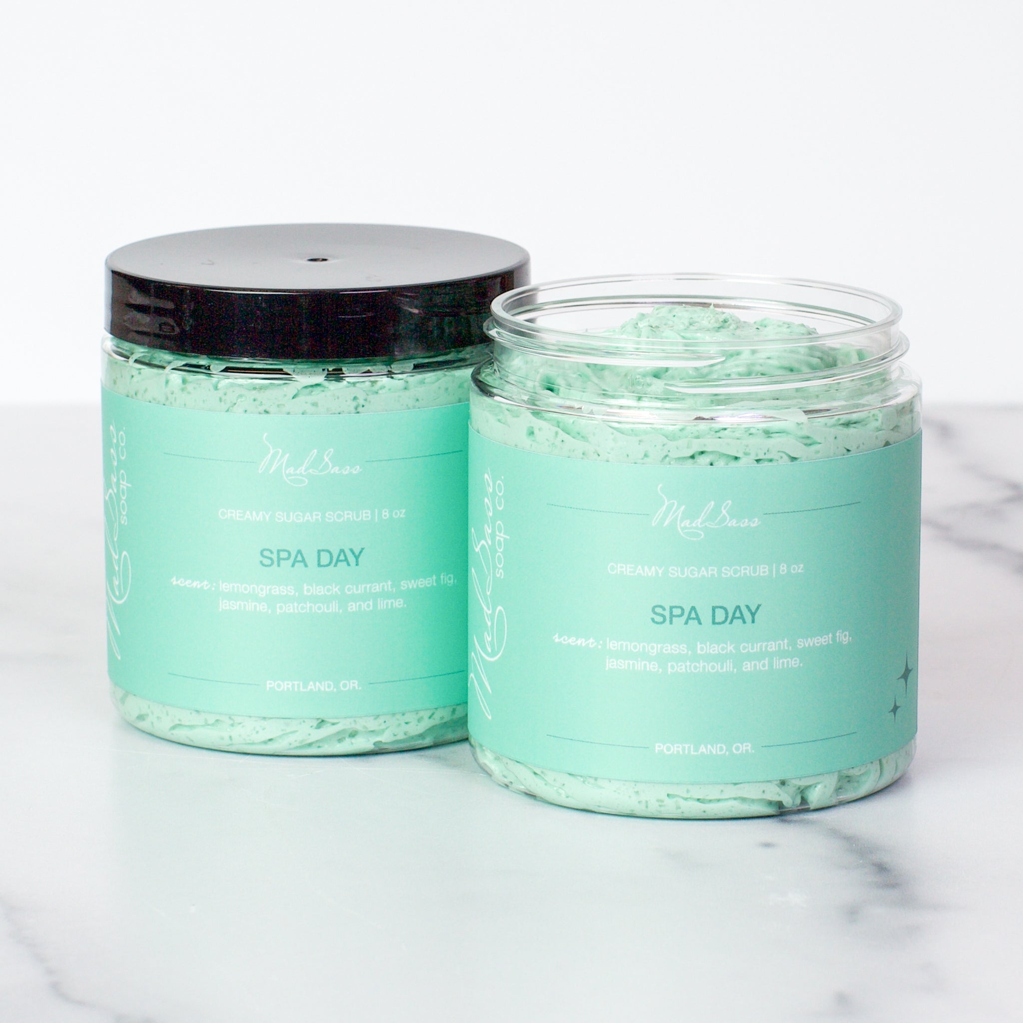 Two containers of Spa Day Creamy Sugar Scrubs on a white background. Spa Day is a light teal scrub in a clear tub.