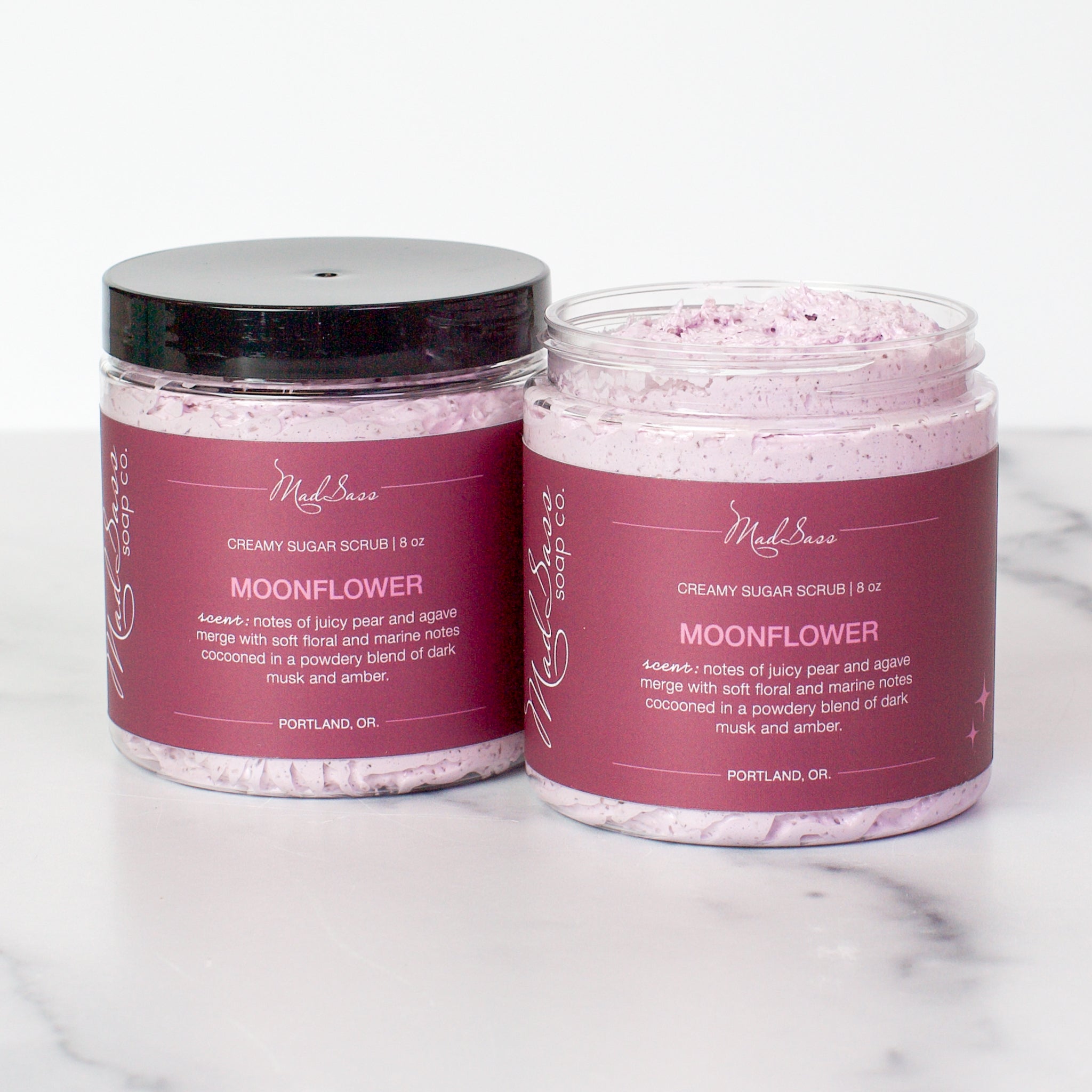 Two containers of Moonflower Creamy Sugar Scrubs on a white background. Moonflower is a light berry scrub in a clear tub with a black lid.