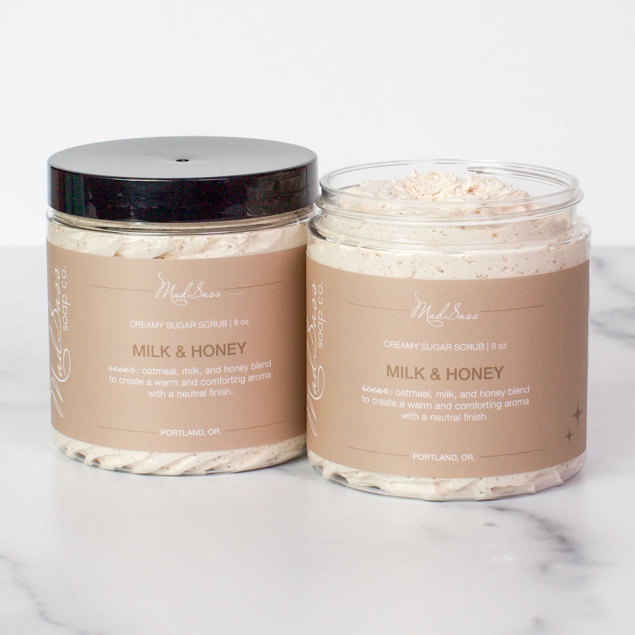 Two containers of Milk & Honey Creamy Sugar Scrubs on a white background. Milk & Honey is a light tan scrub in a clear tub with a black lid.