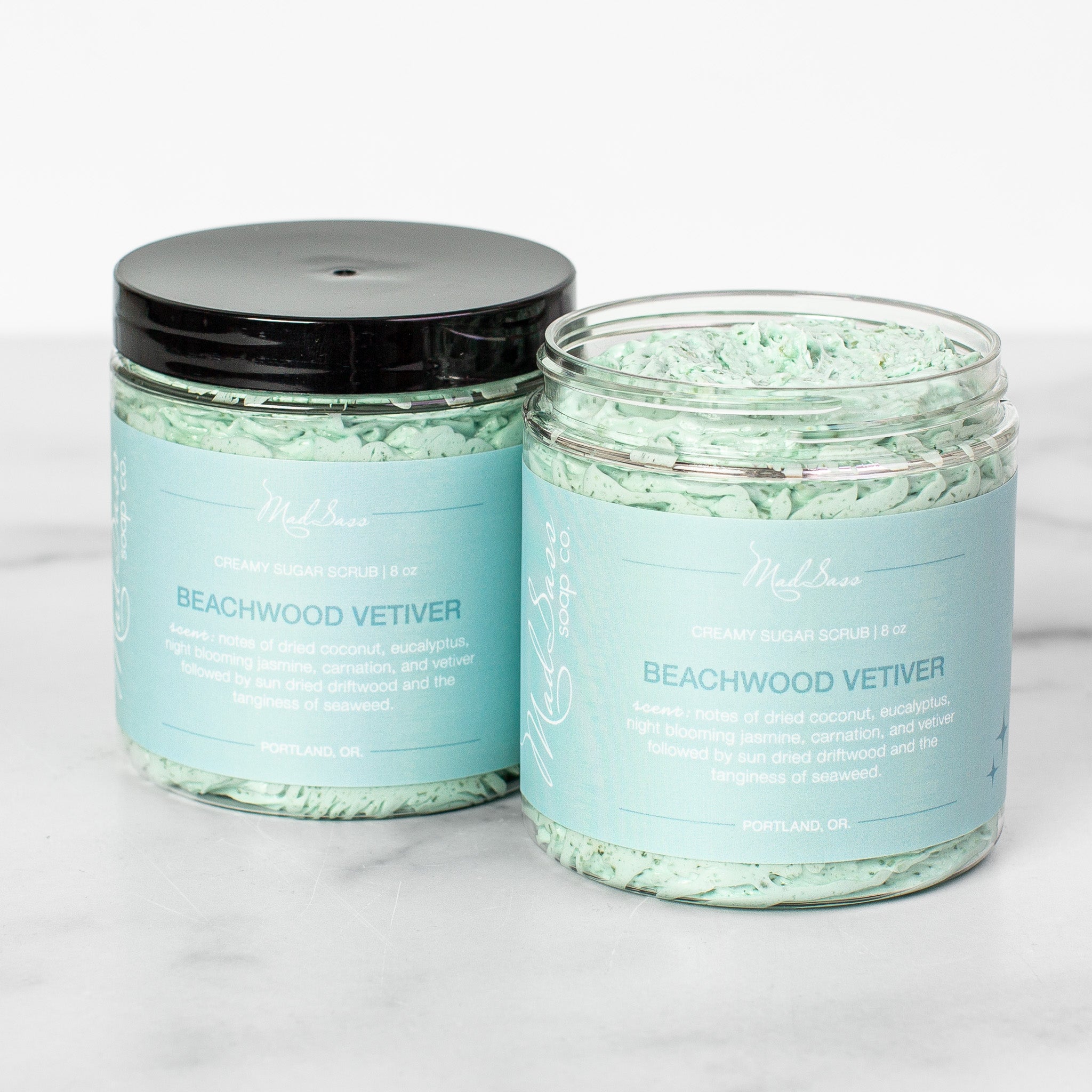 Two containers of Beachwood Vetiver Creamy Sugar Scrubs on a white background. Beachwood Vetiver is a light blue scrub in a clear tub with a black lid.