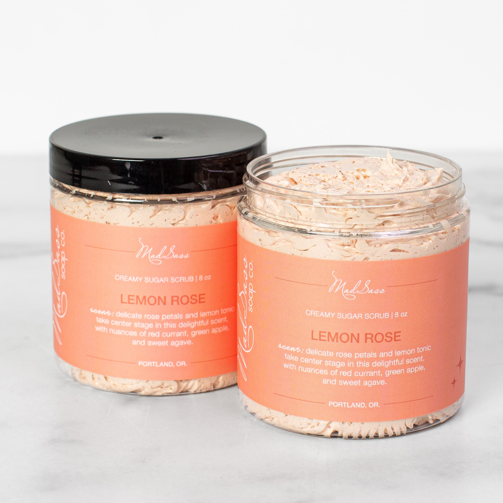 Two containers of Lemon Rose Creamy Sugar Scrubs on a white background. Lemon Rose is a light pink scrub in a clear tub with a black lid.