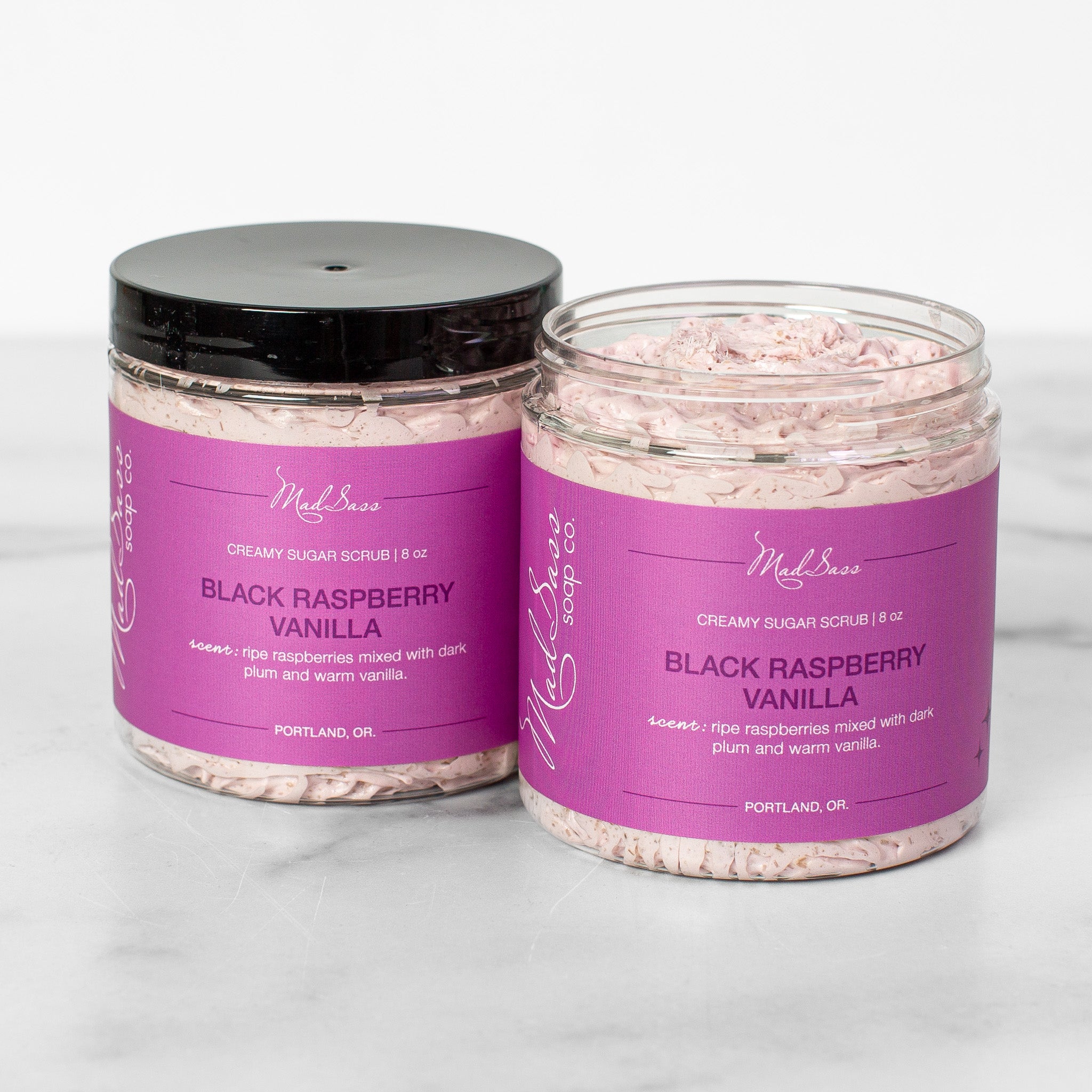 Two containers of Black Raspberry Vanilla Creamy Sugar Scrubs on a white background. Black Raspberry Vanilla is a light purple scrub in a clear tub with a black lid.