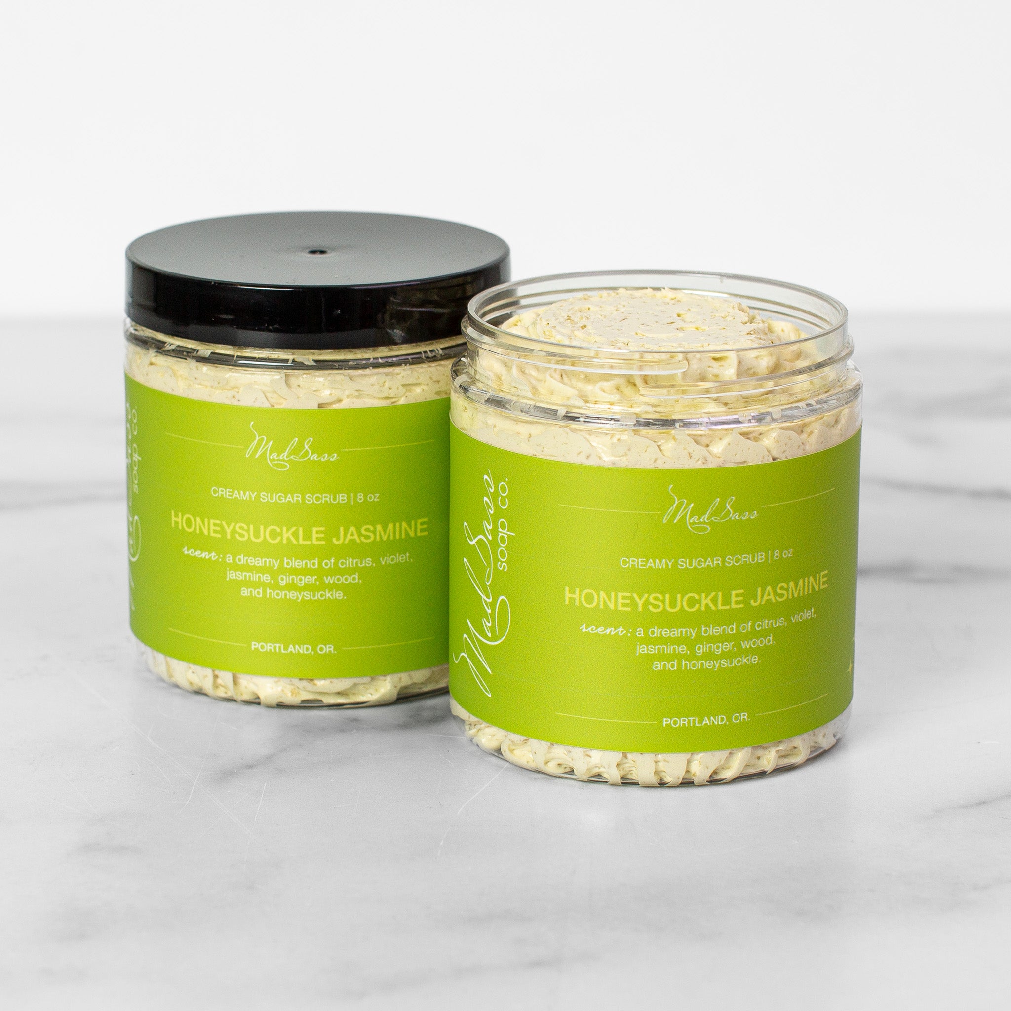 Two containers of Honeysuckle Jasmine Creamy Sugar Scrubs on a white background. Honeysuckle Jasmine is a light green scrub in a clear tub with a black lid.