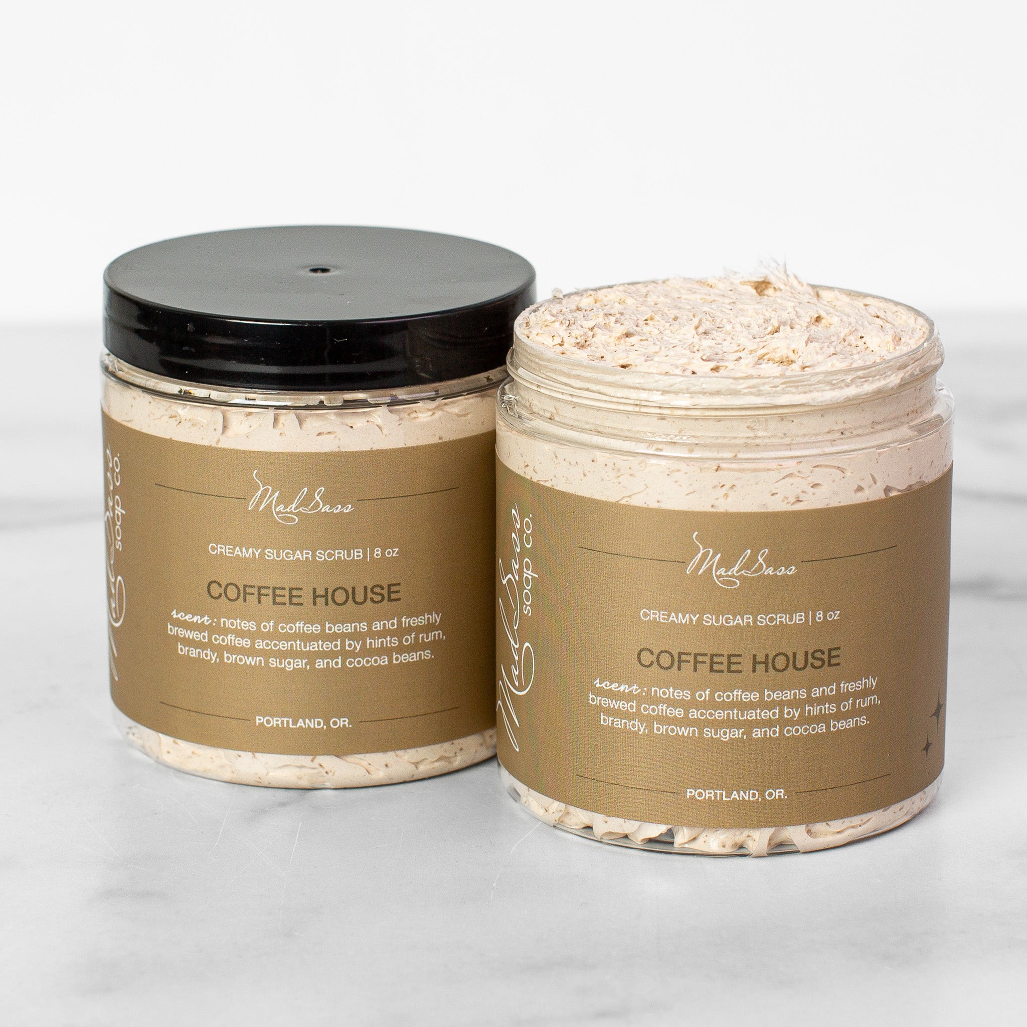Two containers of Coffee House Sugar Scrubs on a white background. Coffee House is a light tan scrub in a clear tub with a black lid.