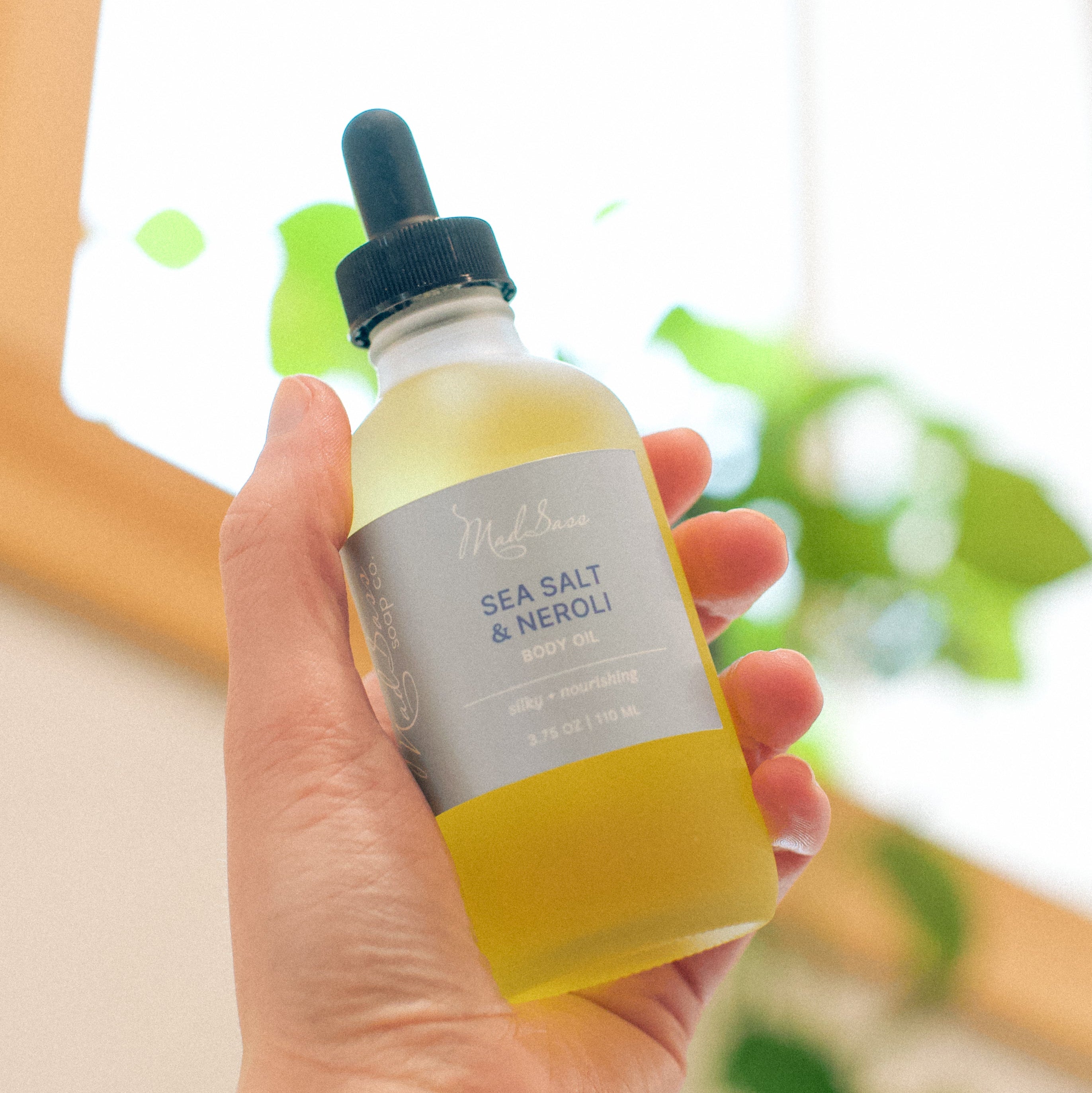 Hand holding the premium Sea Salt & Neroli - Body Oil by Mad Sass Soap Co, emphasizing its luxurious formula as a skin nourishing body oil, surrounded by a lush green ambiance.
