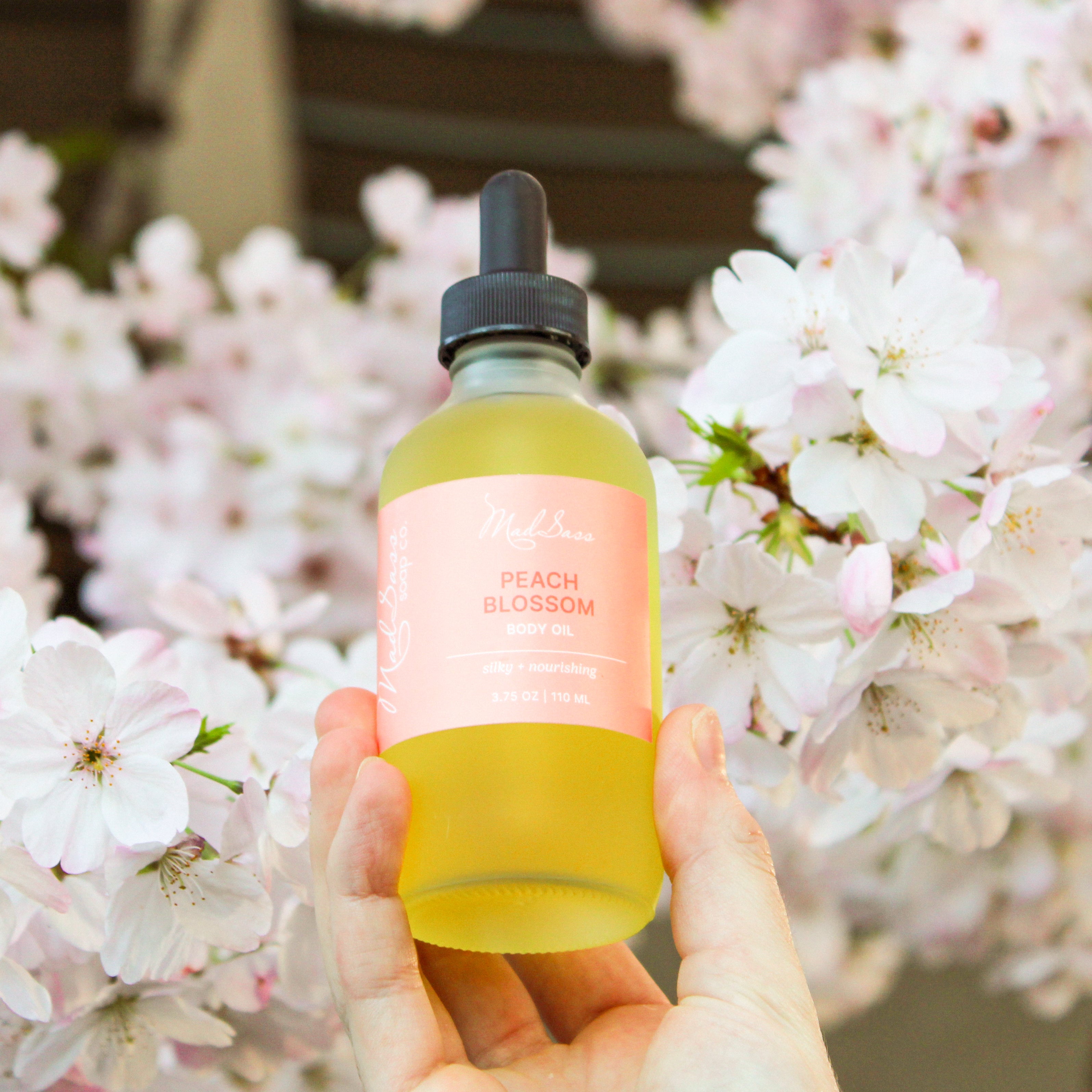 Hand holding a bottle of Peach Blossom skin nourishing body oil by Mad Sass Soap Co against a backdrop of blooming flowers, highlighting its natural and nourishing qualities.