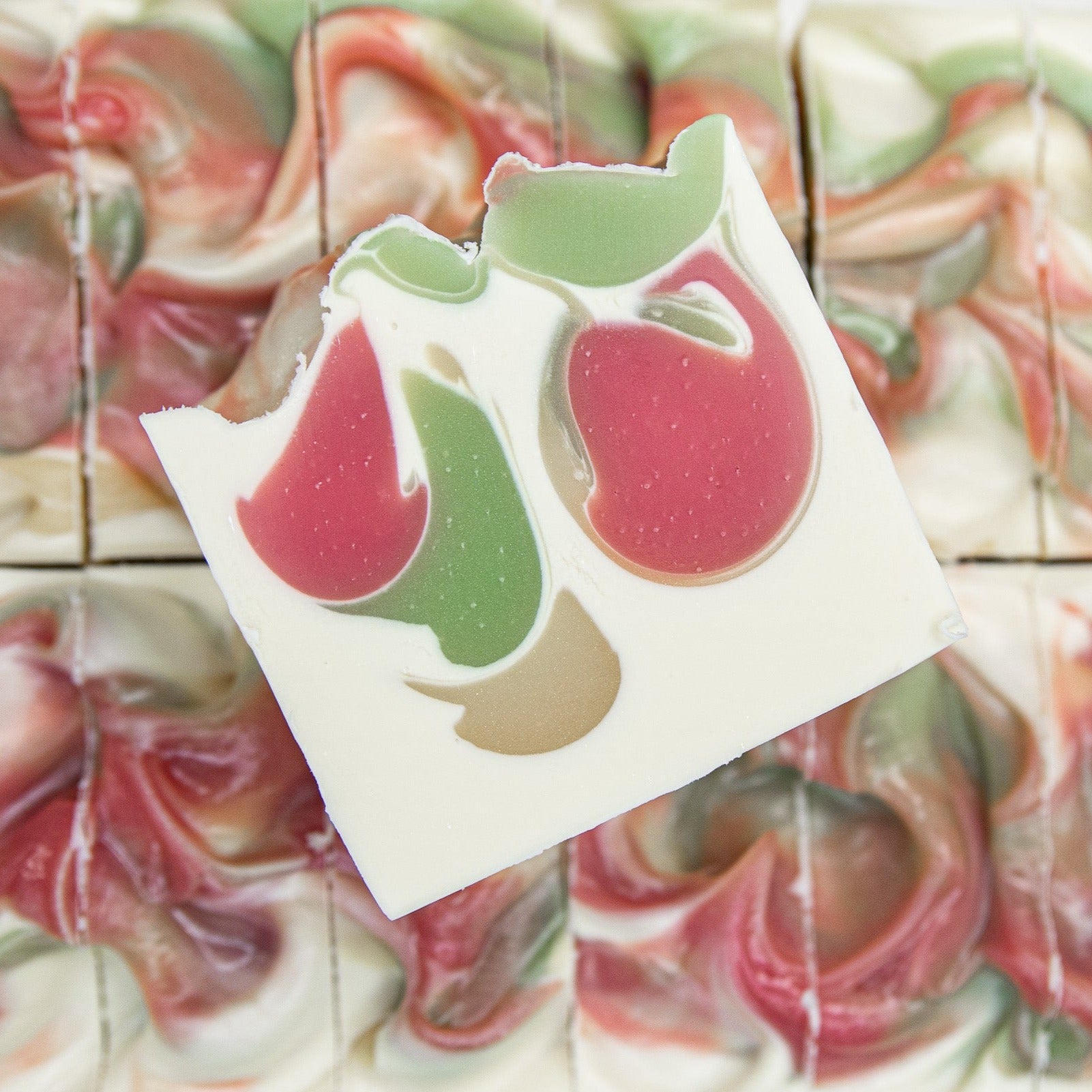 One bar of Apple Sage soap laying on the top of two rows of Apple Sage soap. Apple Sage is an off-white soap with a pink-red, green, and yellow drop swirl design.
